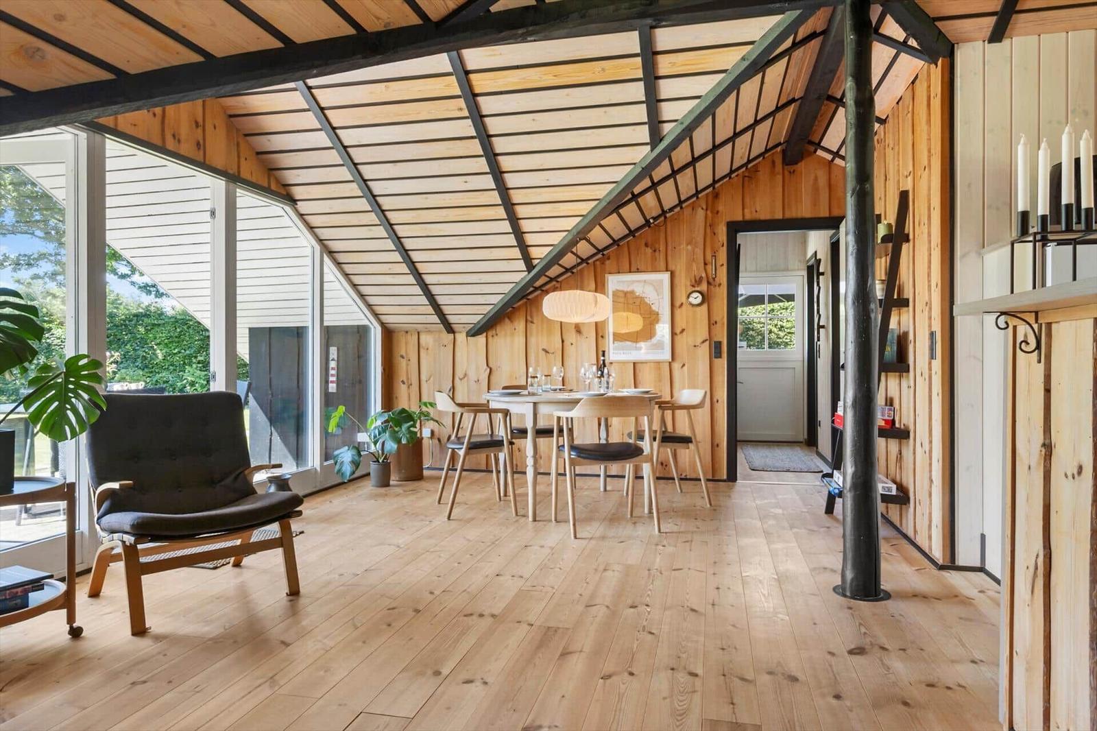 Ein Raum mit Holzdecke, -wänden und -boden. Es gibt einen Tisch mit Stühlen, einen Sessel und große Fenster.
