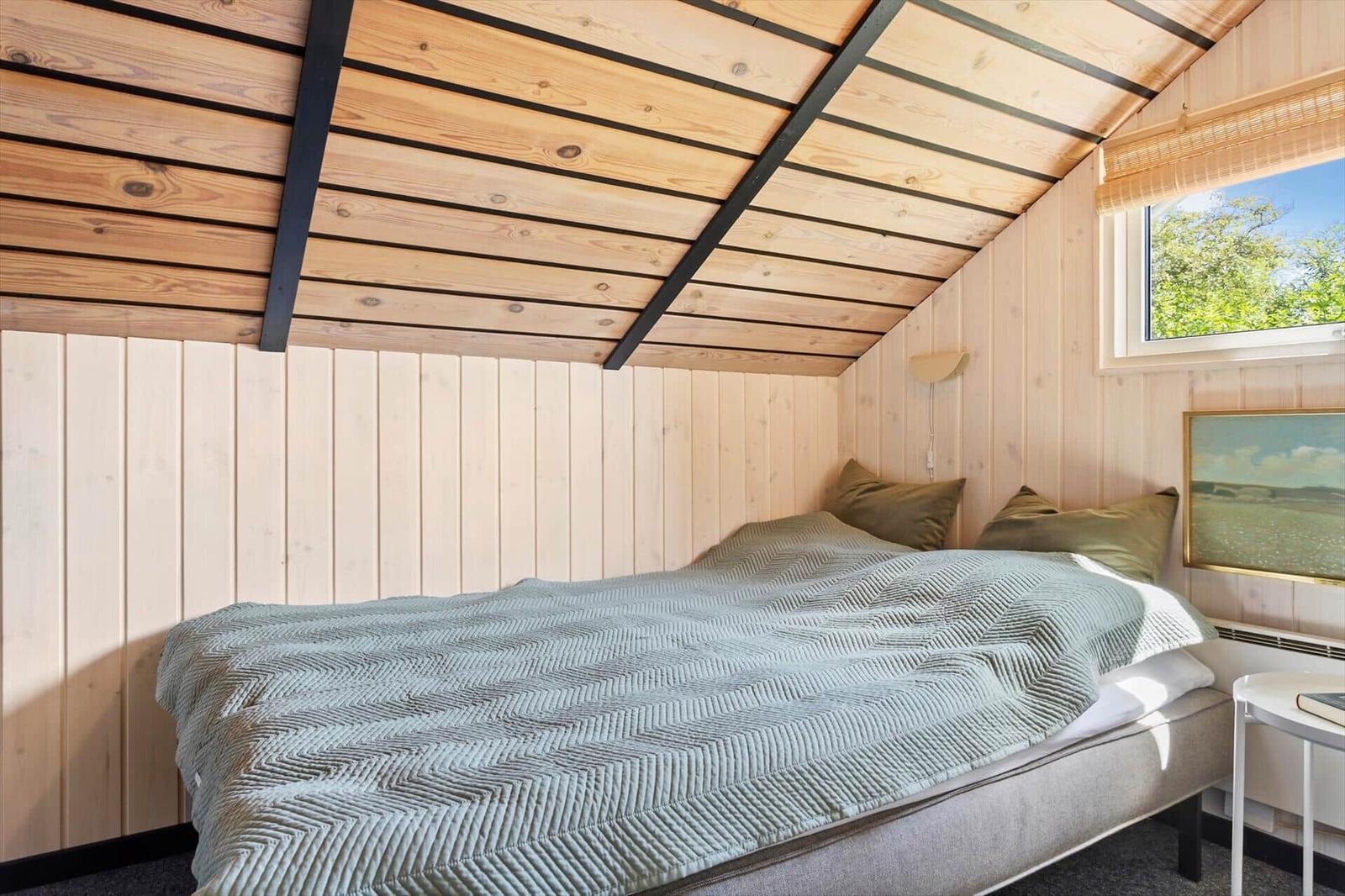 Schlafzimmer mit Bett, Holzwand und Dachbodenstruktur. Fenster mit Blick auf Grün.