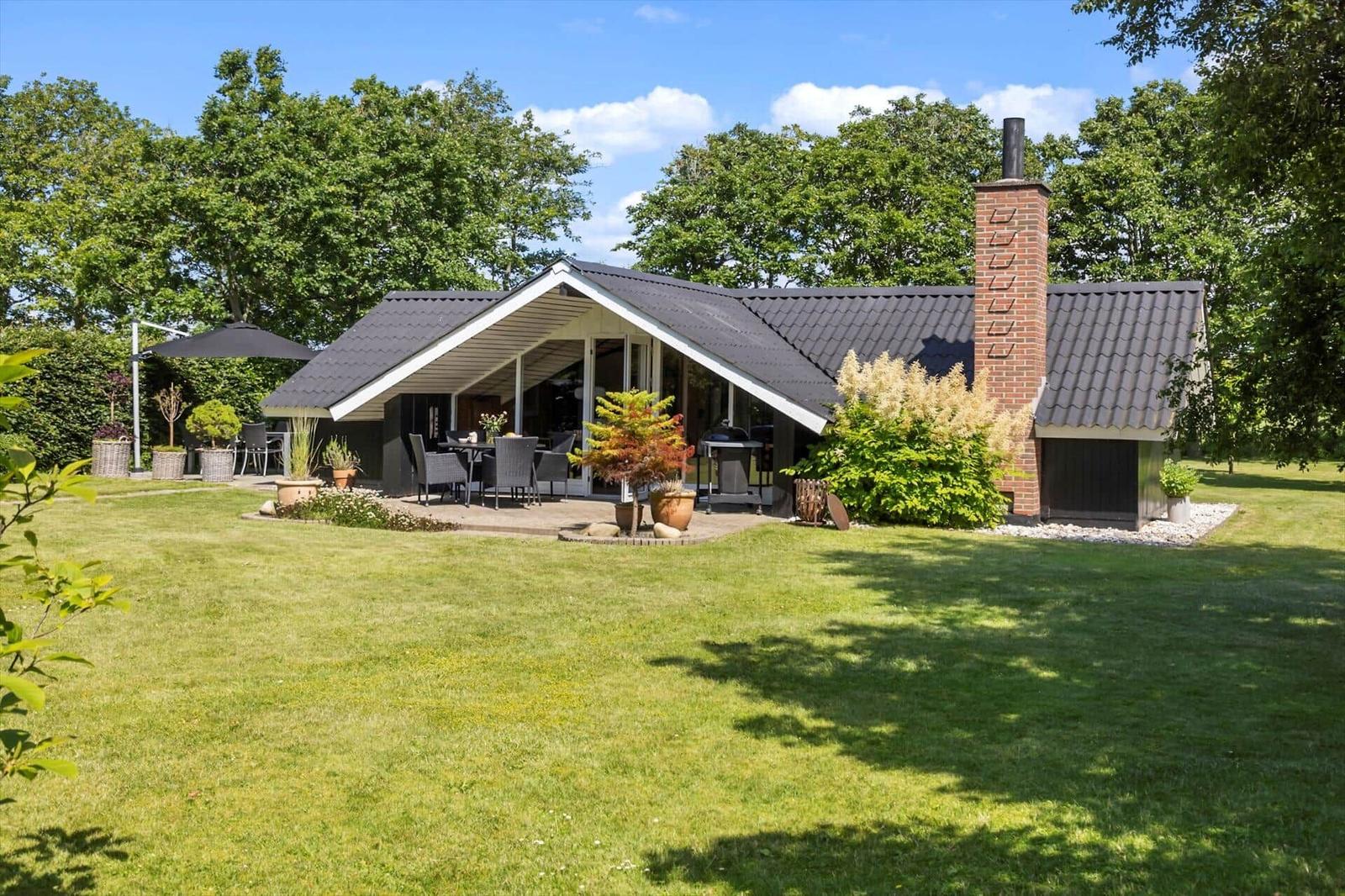 Moderne Villa mit Terrasse, Grill und Garten. Roter Schornstein und grüne Bäume im Hintergrund.
