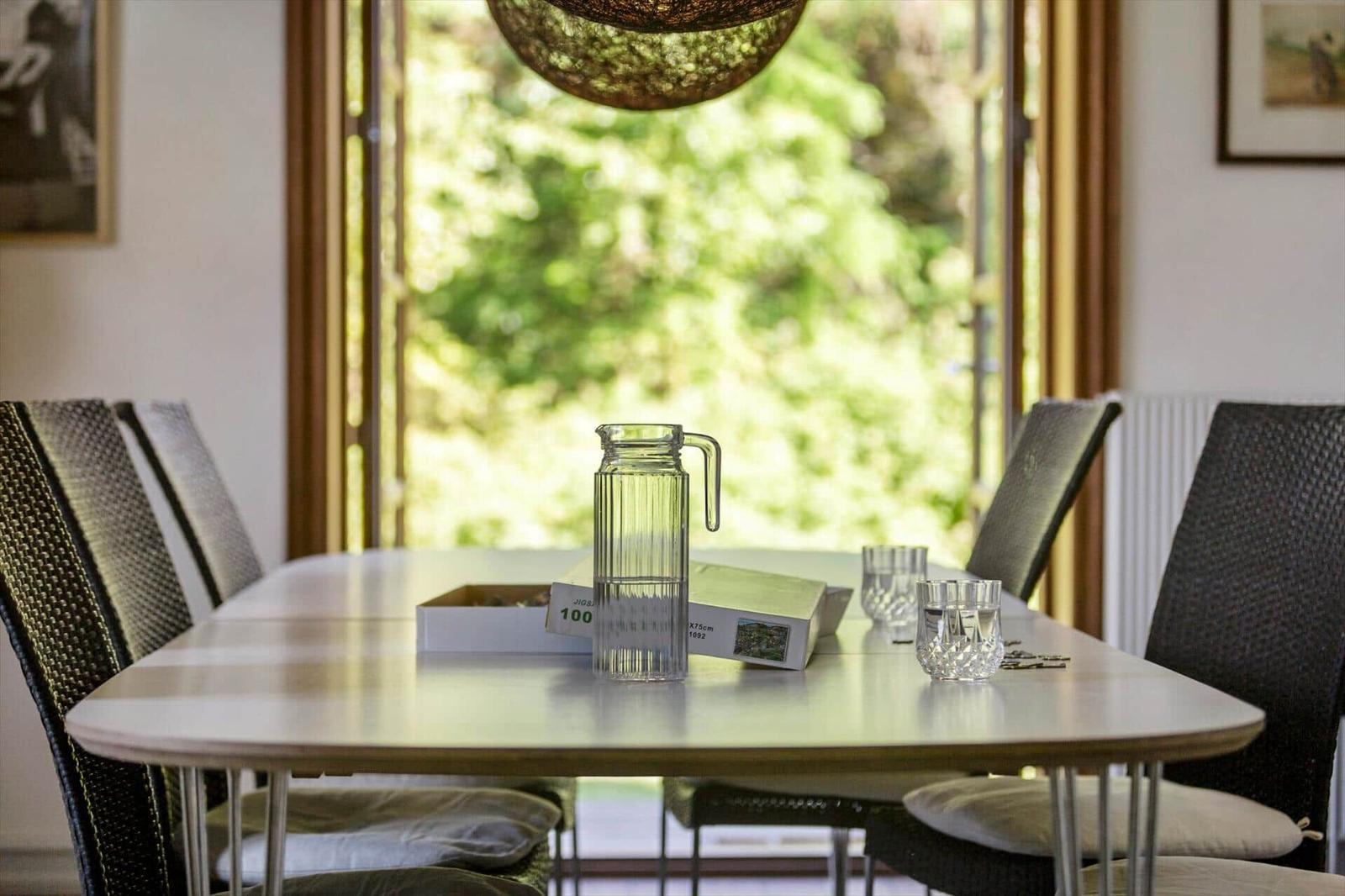 Esstisch mit Stühlen und Wasserglas vor großem Fenster mit Blick auf Garten.
