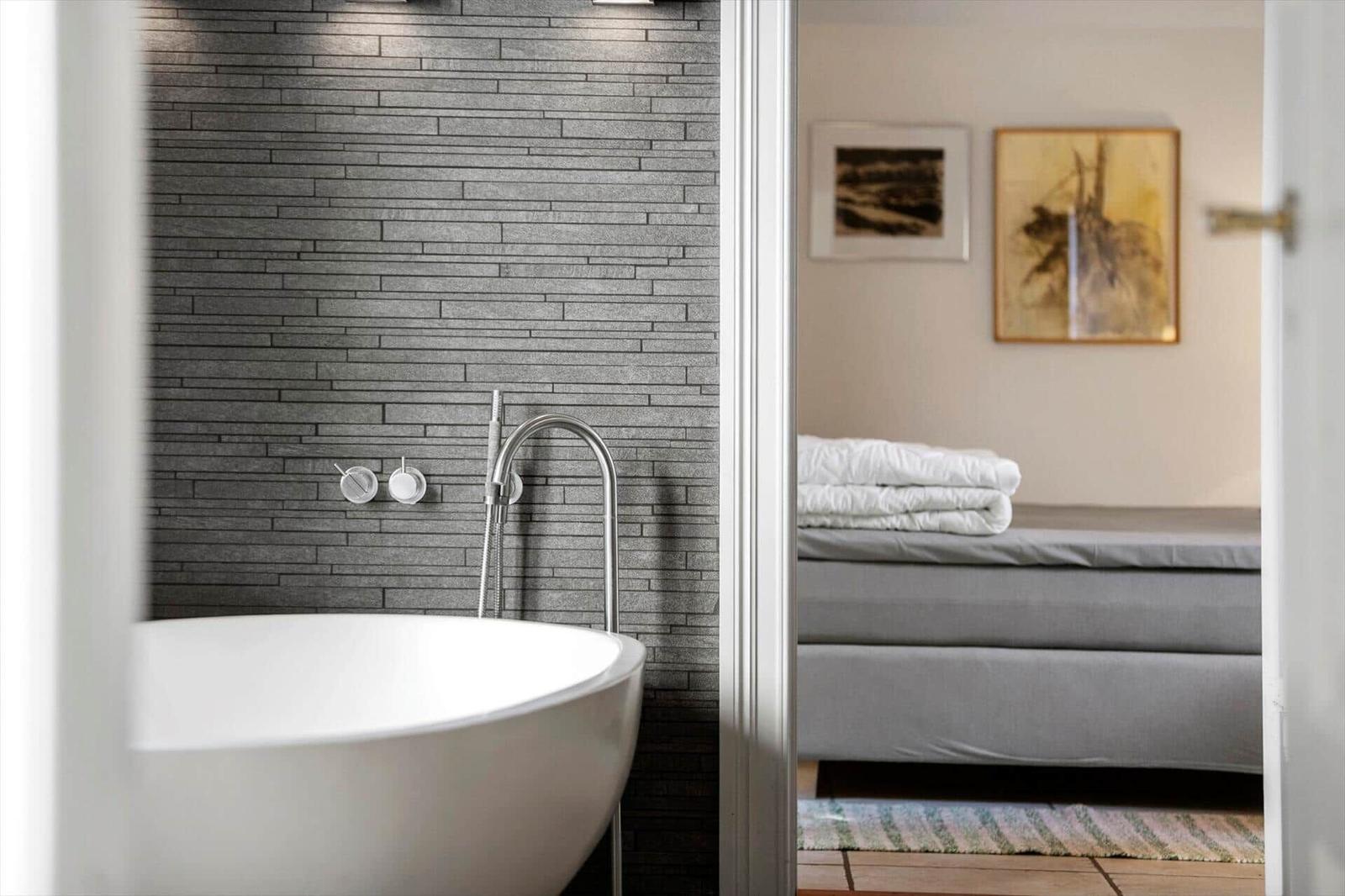 Bad mit freistehender Badewanne und grauer Wand. Im Hintergrund Schlafzimmer mit Bett und Kunstwerken.