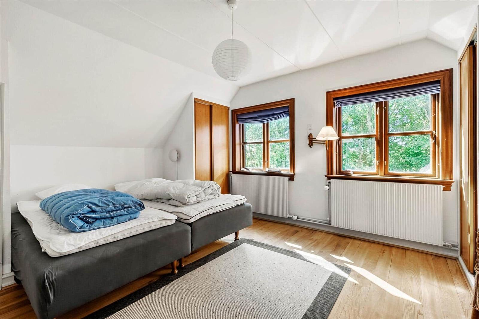 Schlafzimmer mit Doppelbett, Holzboden und zwei Fenstern mit Blick auf Bäume.