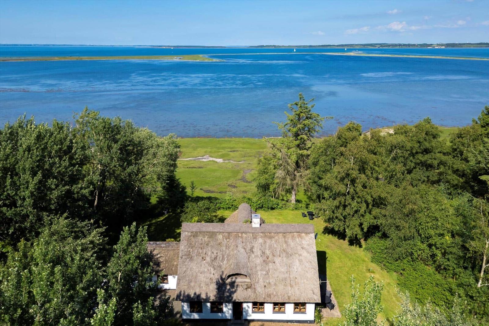Haus mit Strohdach umgeben von Bäumen und Gras, Blick auf blaues Wasser und Insel.