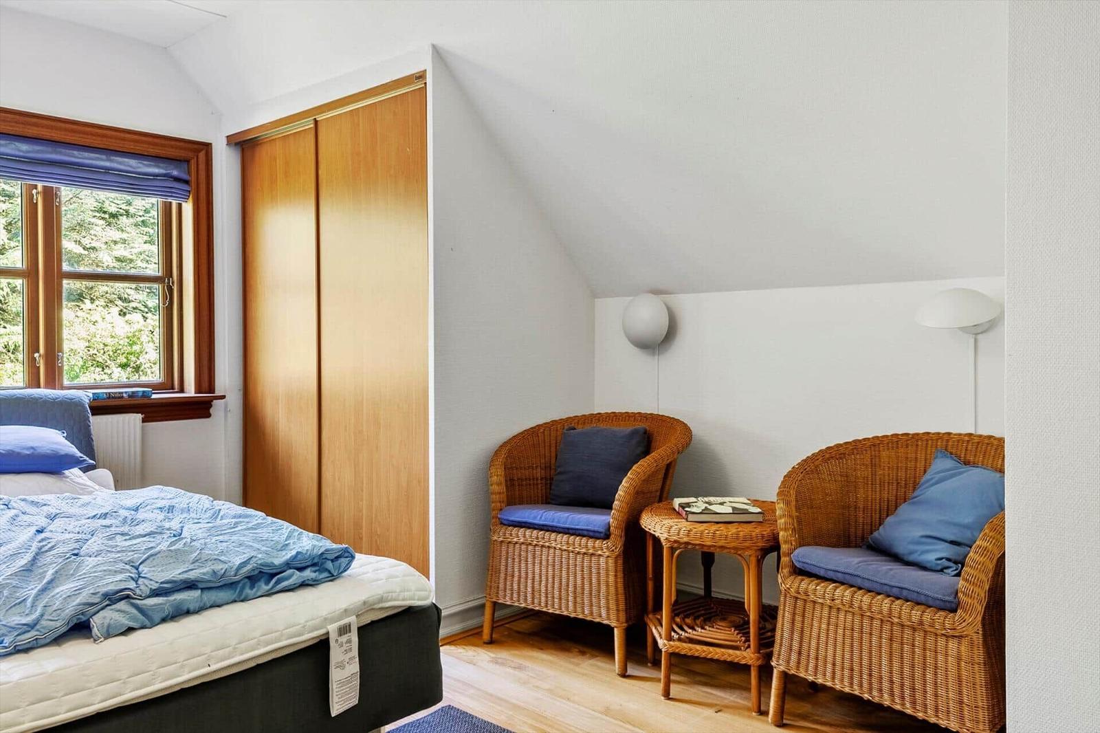 Schlafzimmer mit Bett, zwei Rattanstühlen und einem Fenster mit Blick auf die Natur.