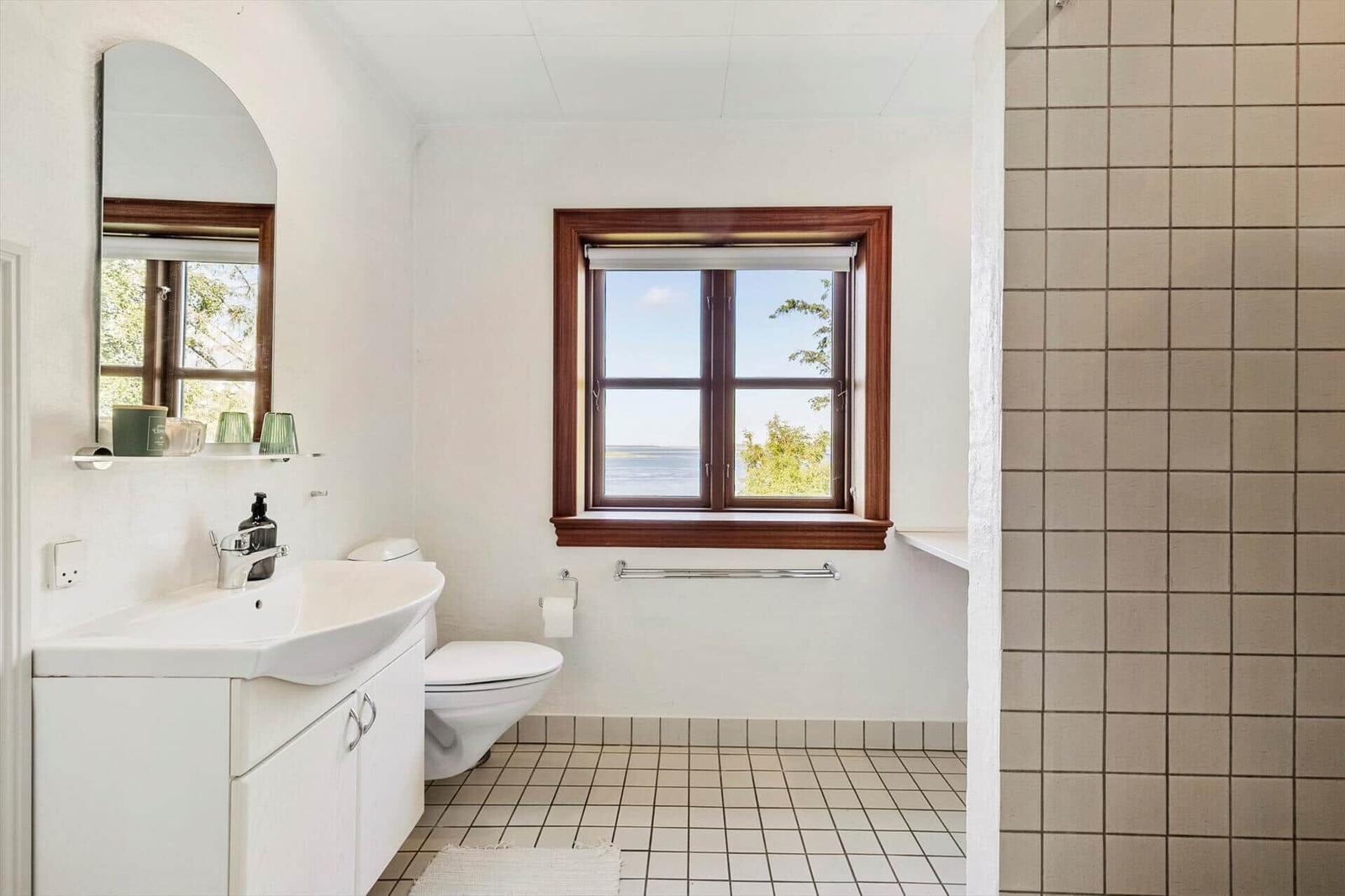 Badezimmer mit Waschbecken, Toilette und Dusche. Fenster mit Blick auf Wasser.