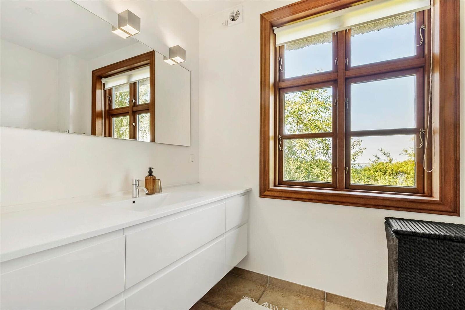 Badezimmer mit Waschbecken, Spiegel und Holzfenster mit Blick auf Bäume.