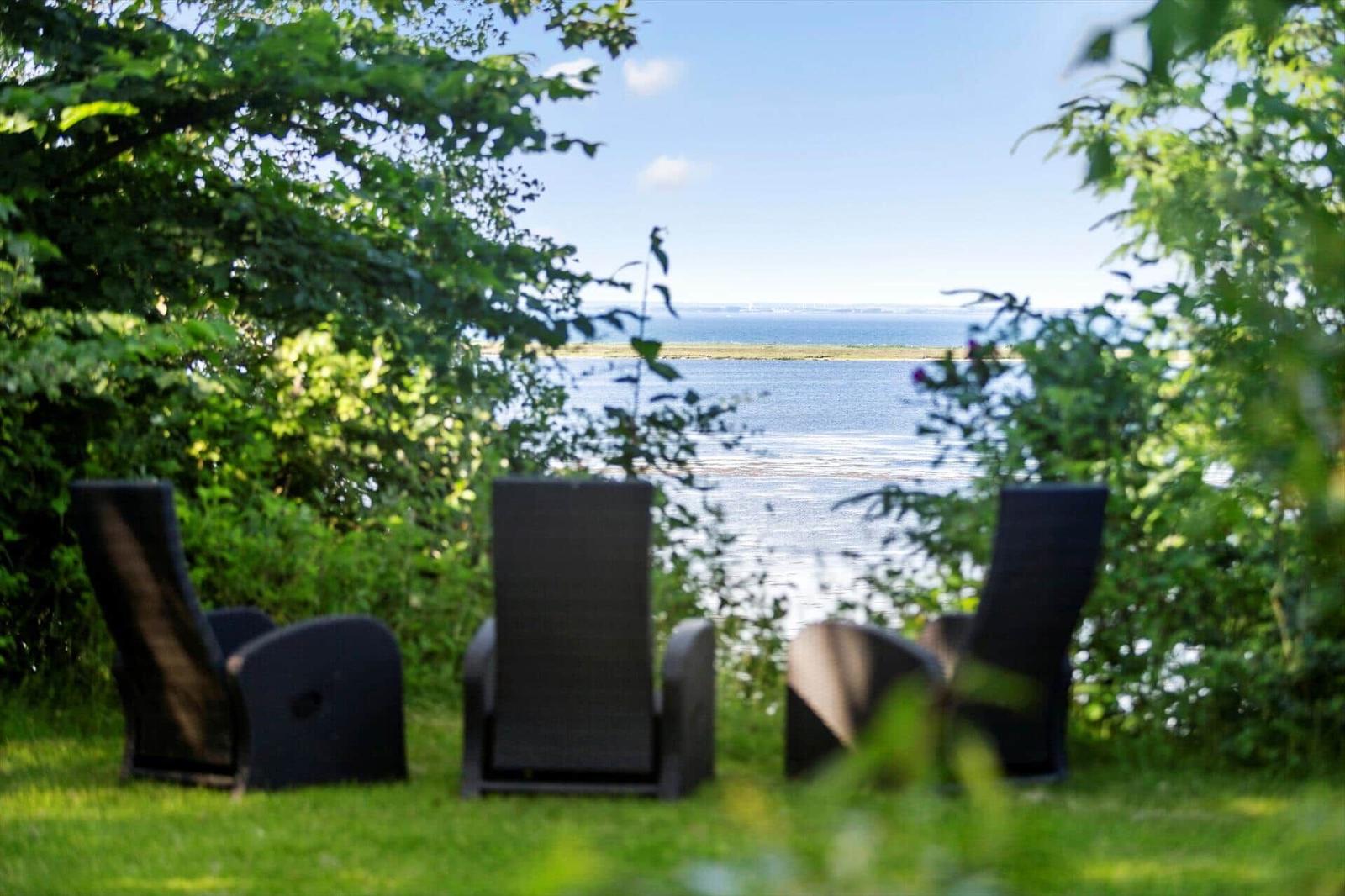 Drei dunkle Liegestühle auf einer Wiese mit Blick auf einen See und einen Strand.