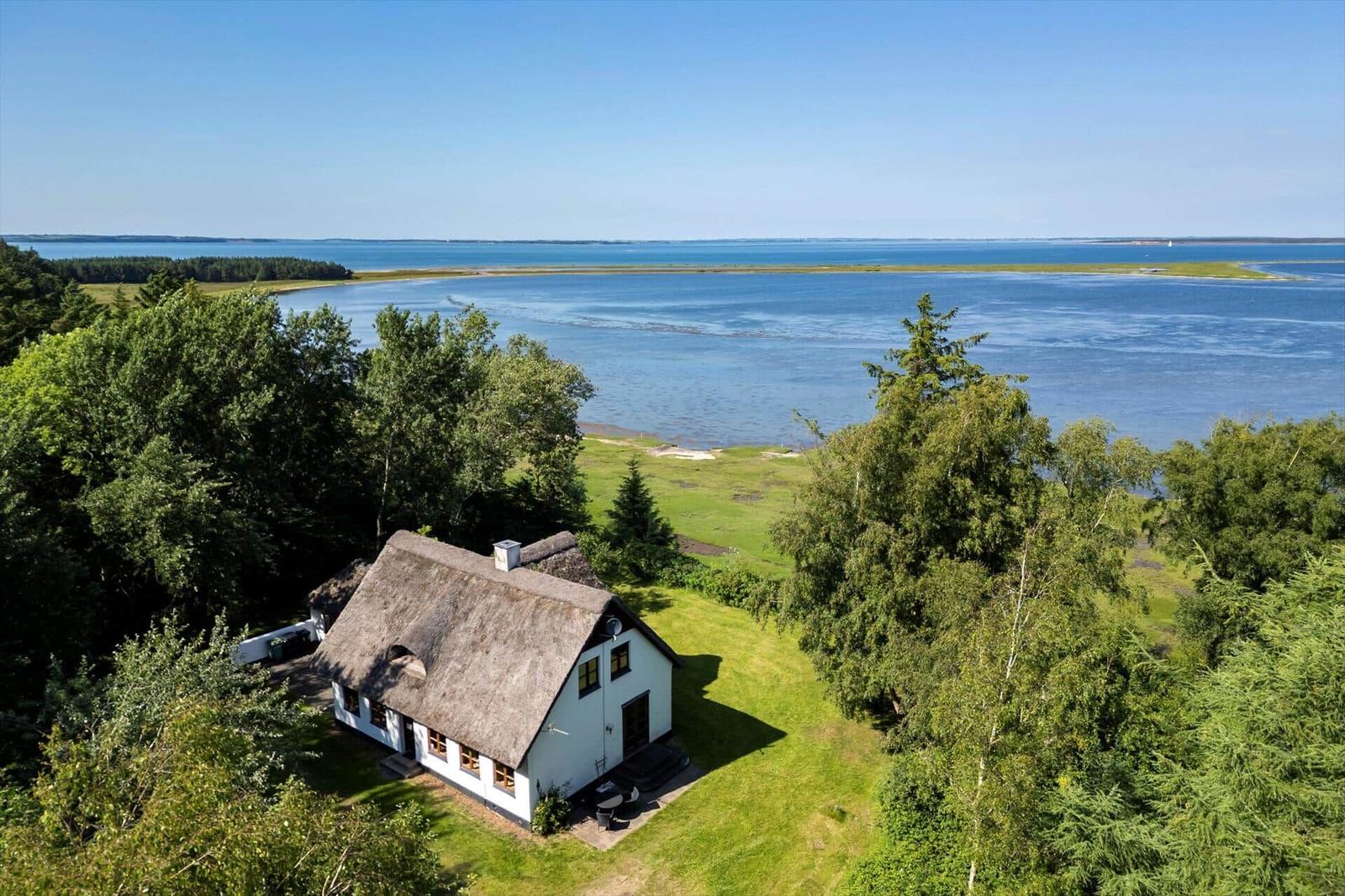 Haus mit Strohdach liegt am Wasser, umgeben von Grün und Bäumen.