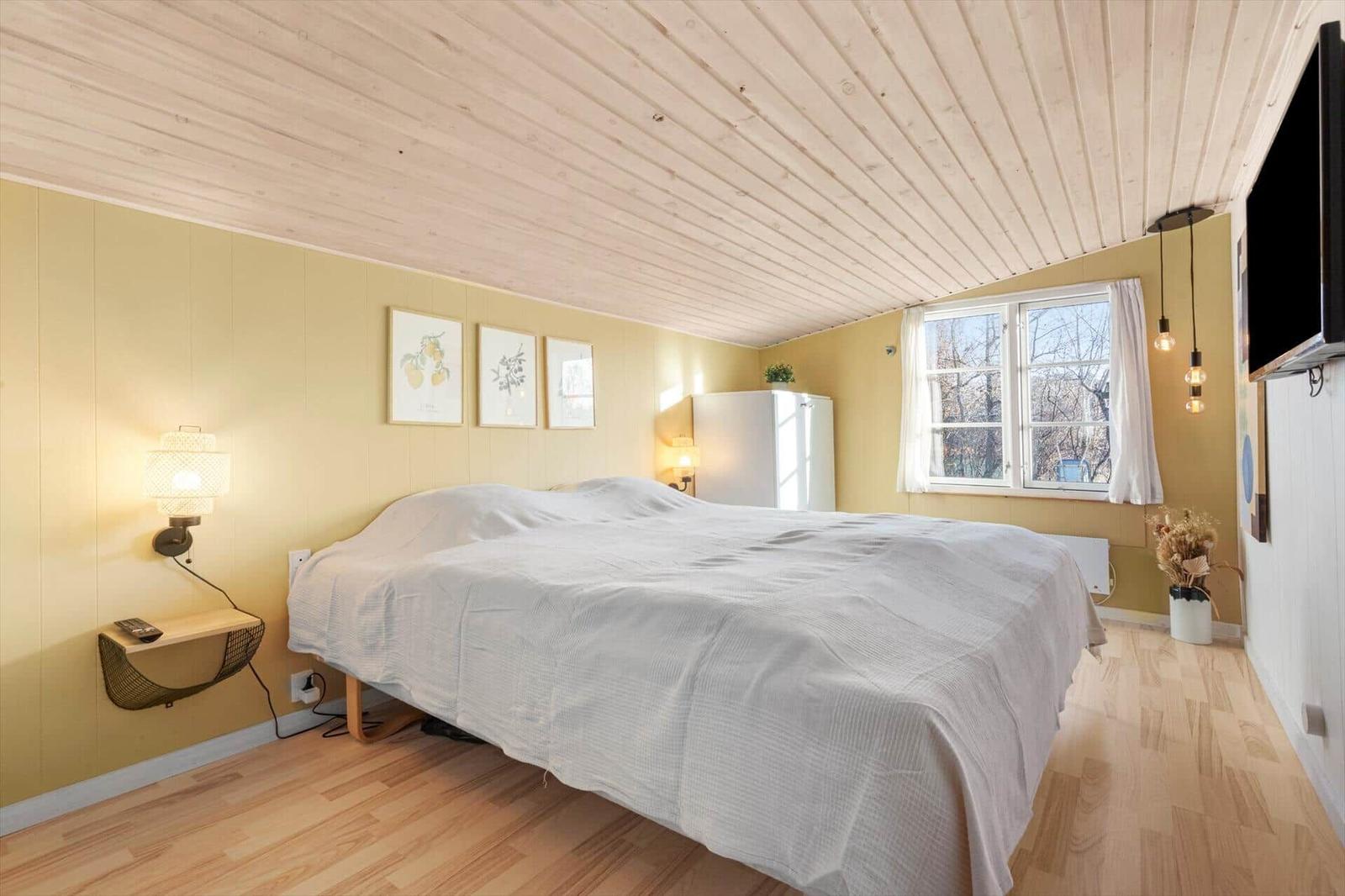 Schlafzimmer mit großem Bett, gelben Wänden und Holzboden. Fenster, TV und Wandlampen.