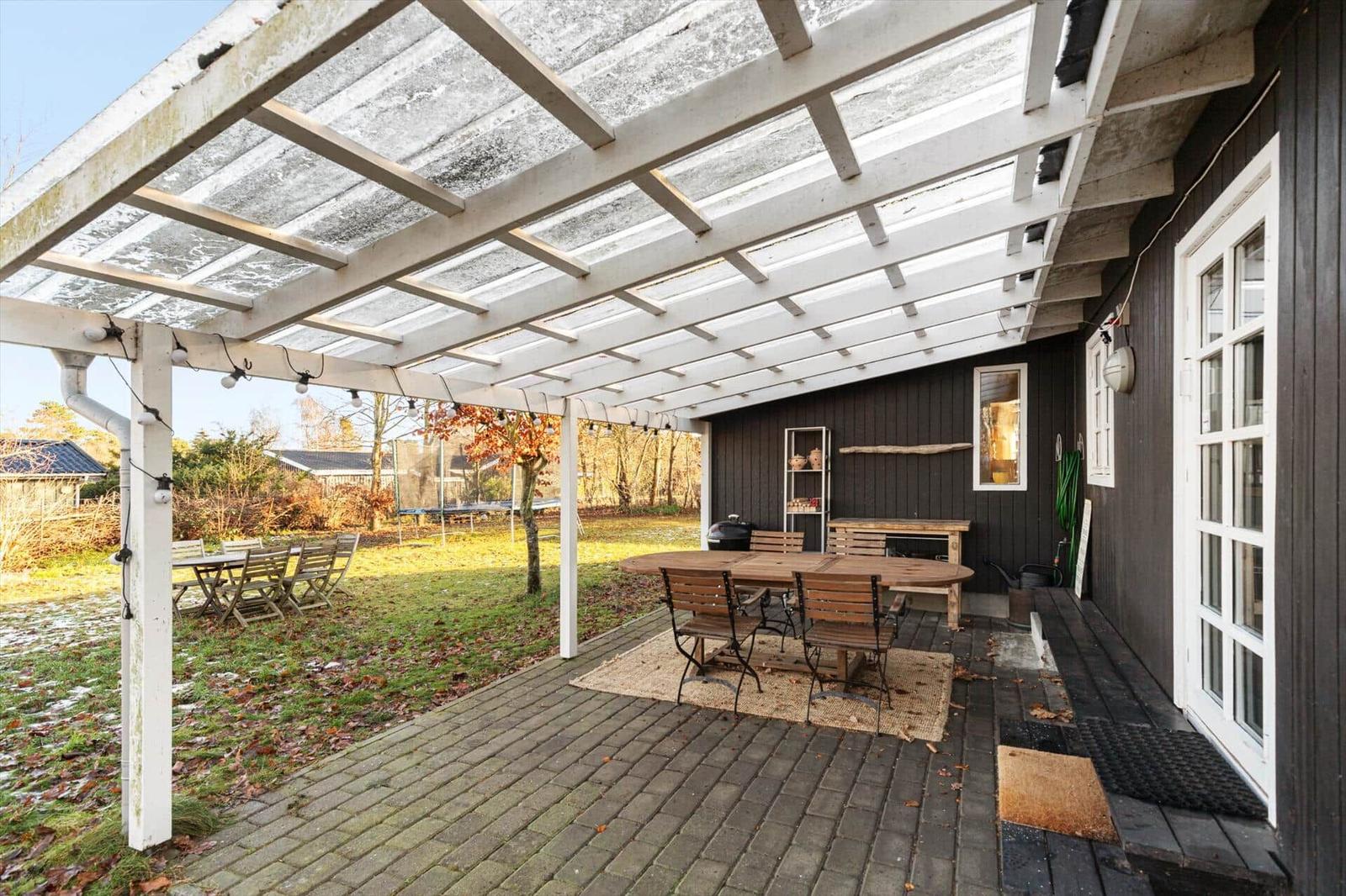 Terrasse mit Holztisch, Stühlen und Dachkonstruktion. Hintergrund: Garten und Hauswand.