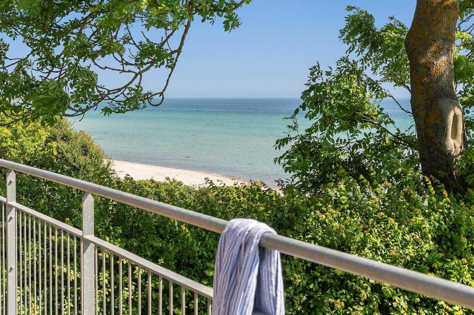 Blick von Balkon auf Strand und Meer durch grüne Bäume und Sträucher.