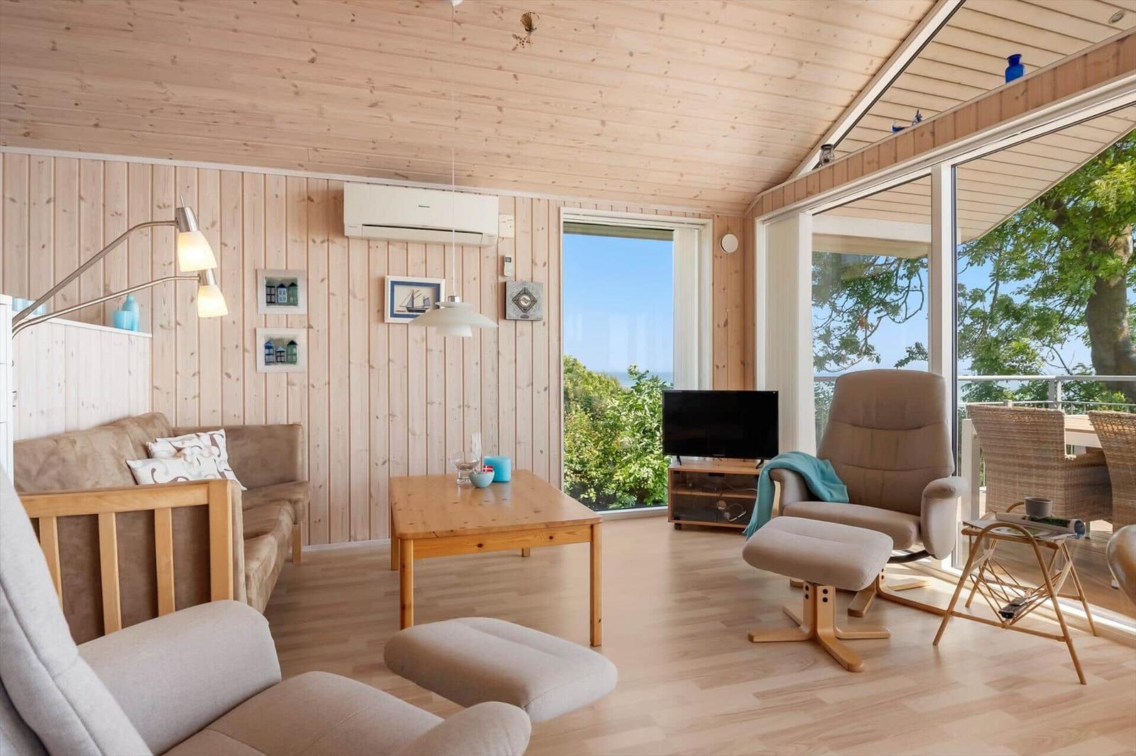 Wohnzimmer mit Holzwand, Sofa, Sessel und Fernseher. Große Fenster mit Blick auf Grün.