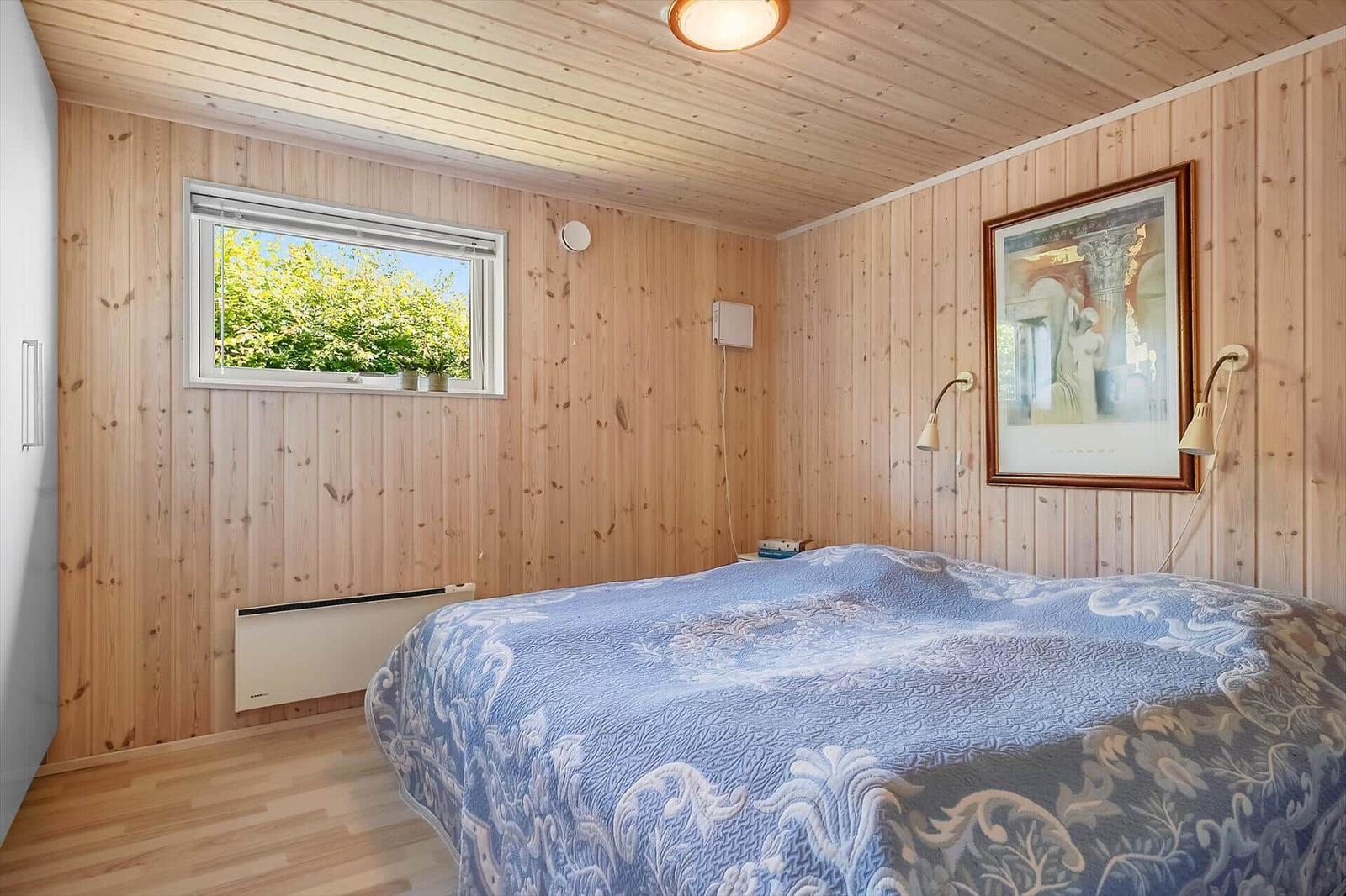 Schlafzimmer mit Holzwandverkleidung, Bett mit blauem Tagesdecke und Fenster mit Blick auf Bäume.