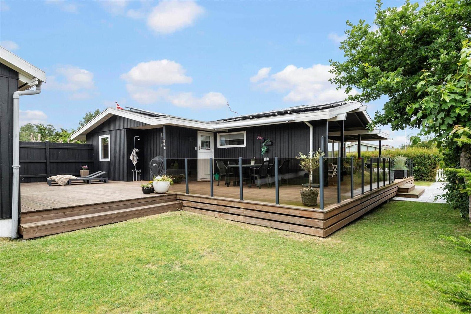 Schwarzes Haus mit großem Holzterrasse und Garten. Außentisch, Liegestühle und Pflanzen.