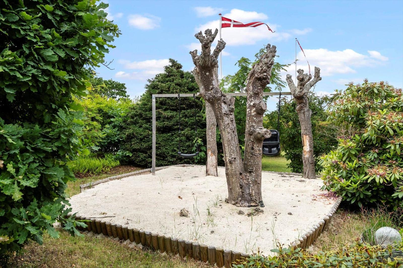 Sandkasten mit Schaukel und dicken Bäumen, daneben Flagge und Grünflächen.