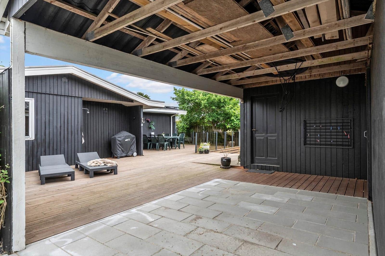 Terrasse mit Liegen, Grill und Tisch unter Holzkonstruktion. Schwarze Fassade und Steinplatten.