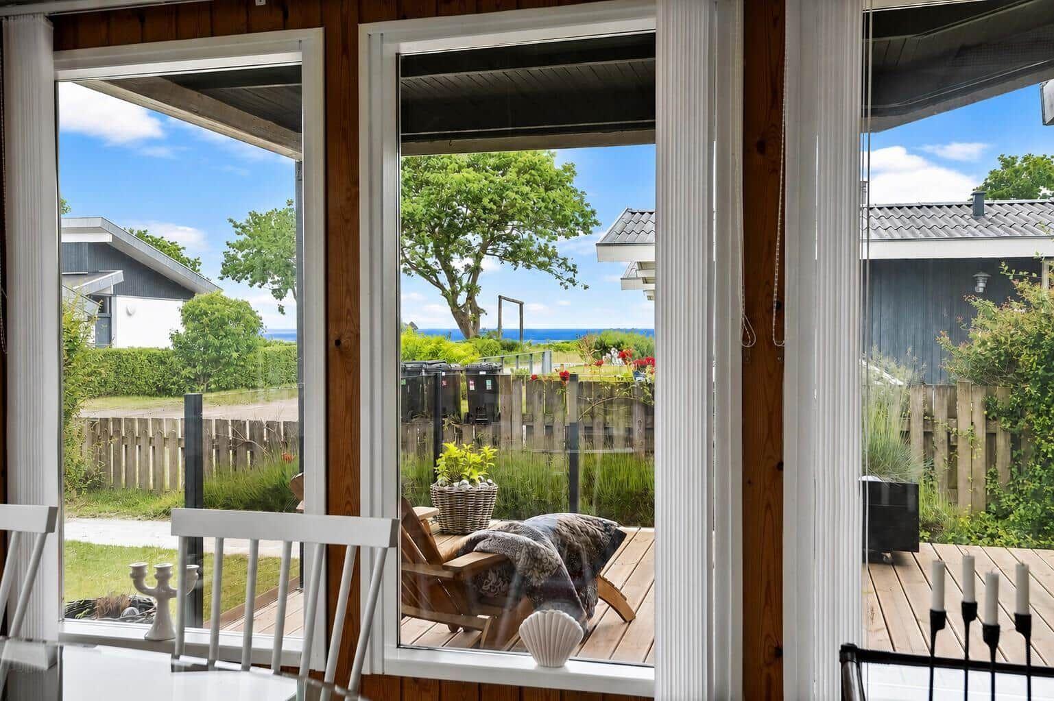 Durch die Fenster ist eine Terrasse mit Sitzgelegenheit und Blick auf Grün und Meer zu sehen.