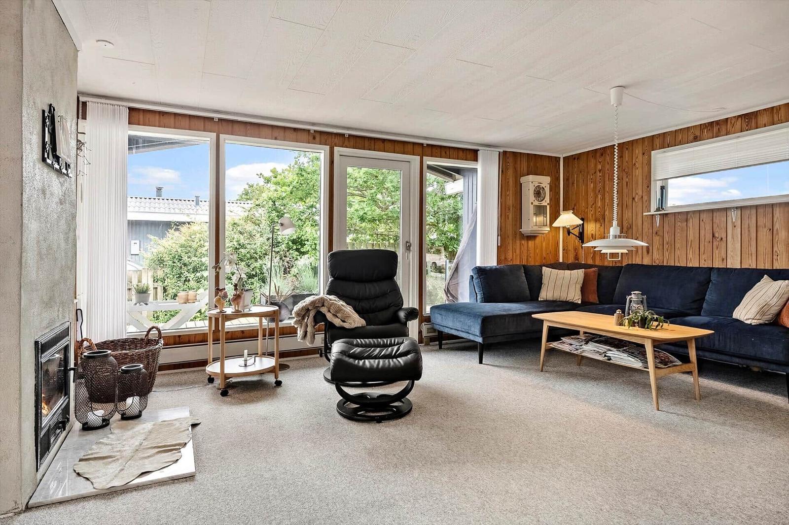 Wohnzimmer mit Sofa, Sessel und Kamin. Große Fensterfront mit Blick auf Garten.
