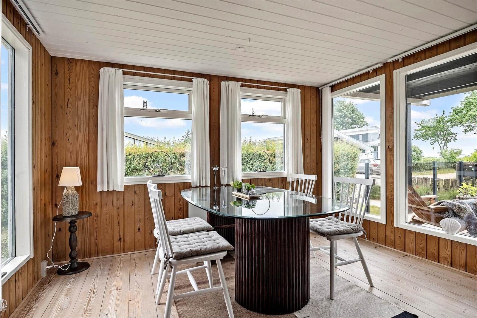 Esszimmer mit Glas-Tisch, weißen Stühlen und Holzwänden. Große Fenster mit Blick nach draußen.