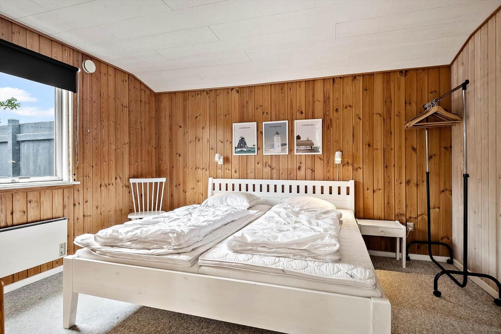 Schlafzimmer mit Holzwand, weißem Bett und Fenster. An der Wand hängen drei Bilder.