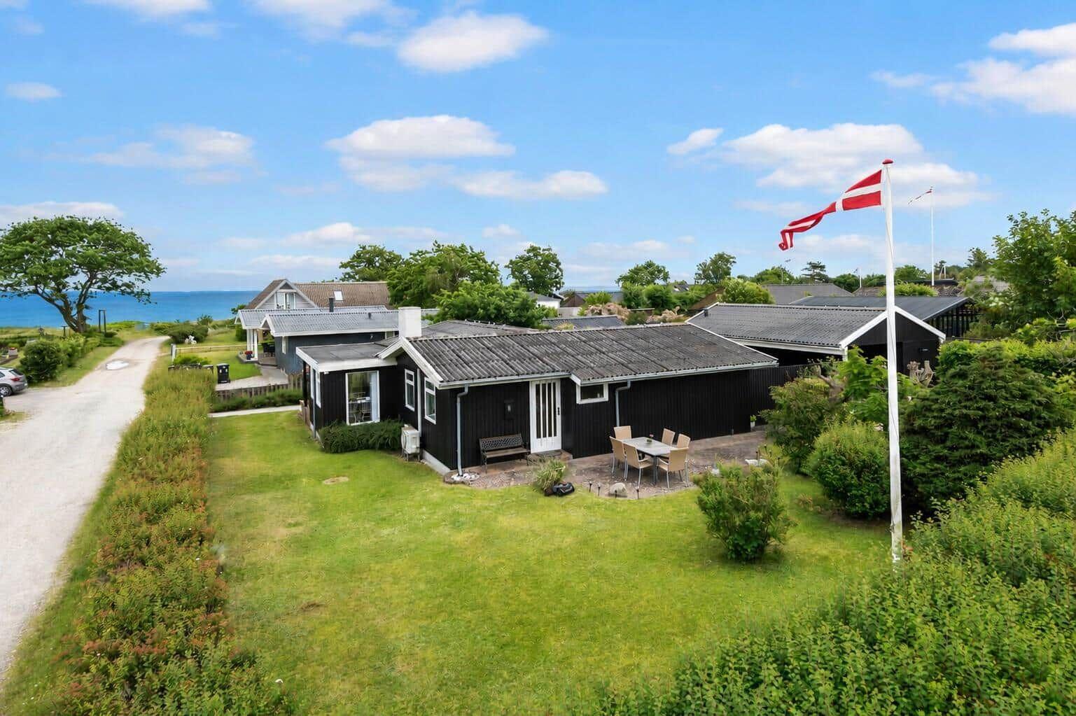 Schwarzes Holzhaus mit Garten, Terrasse und dänischer Flagge. Blick aufs Meer.