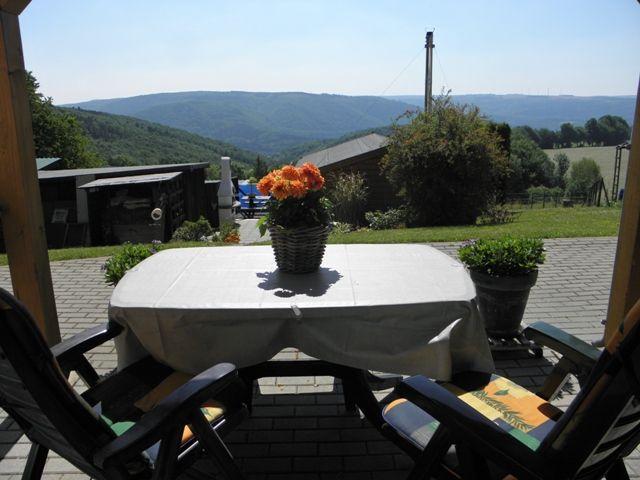 Terrasse mit Tisch, Stühlen und Blumen. Blick auf Hügel und Grünflächen.