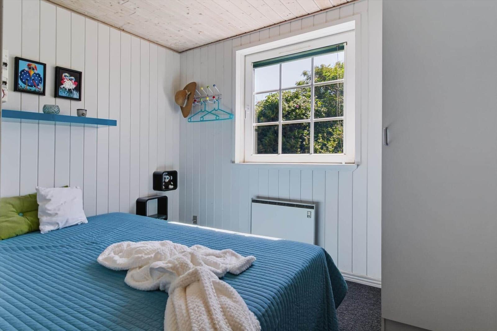 Schlafzimmer mit Bett, Fenster und Wandregal. Wandverkleidung und blaues Bettzeug.