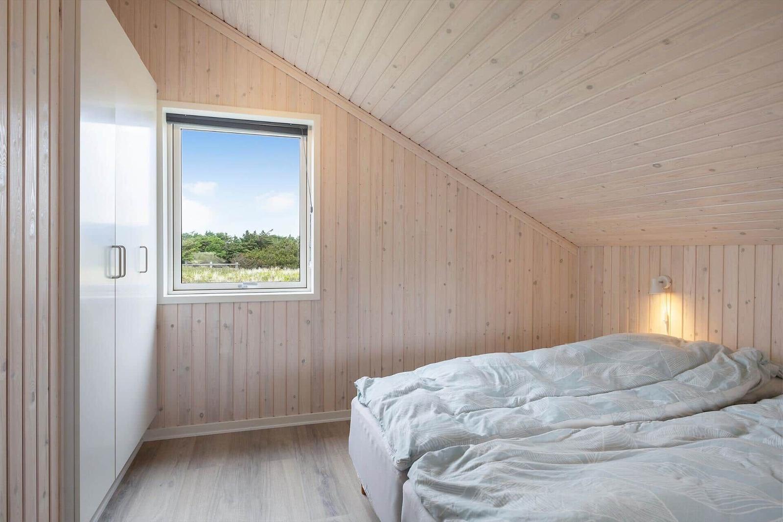 Schlafzimmer mit Holzwand, Bett und Fenster mit Blick auf Grünflächen.