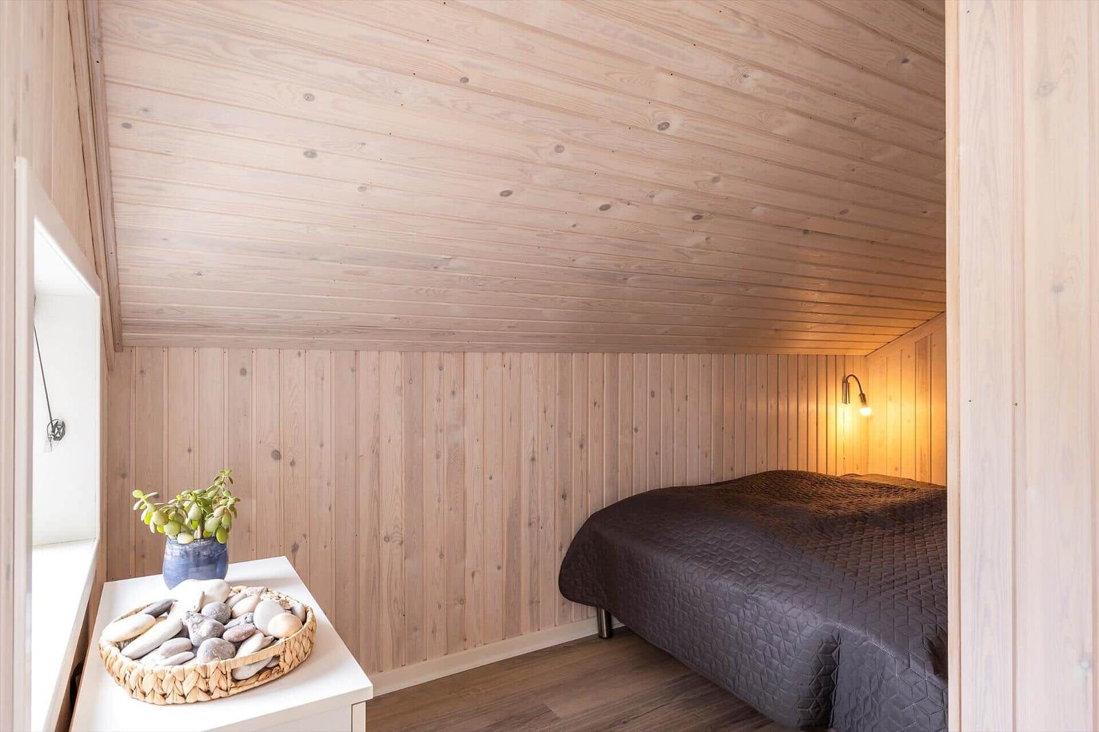 Schlafzimmer mit Holzwand und -decke, Bett mit dunkler Bettwäsche, Nachttisch mit Stein- und Pflanzenarrangement.