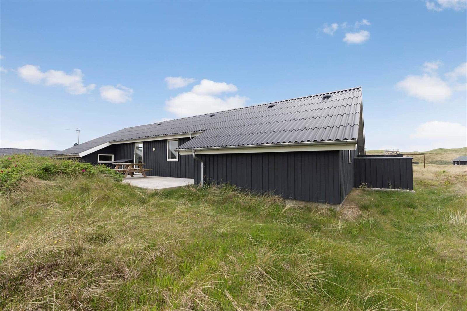 Ein dunkel verkleidetes Ferienhaus mit Terrasse und Picknicktisch in einer grünen Landschaft.