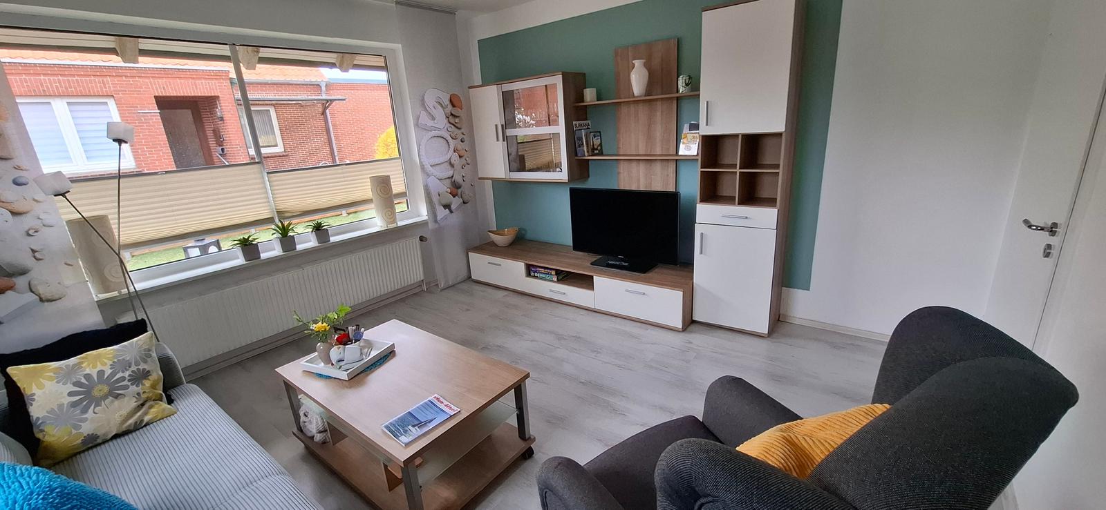 Wohnzimmer mit Sofa, Sessel, Couchtisch und Fernseher. Große Fensterfront mit Blick nach außen.