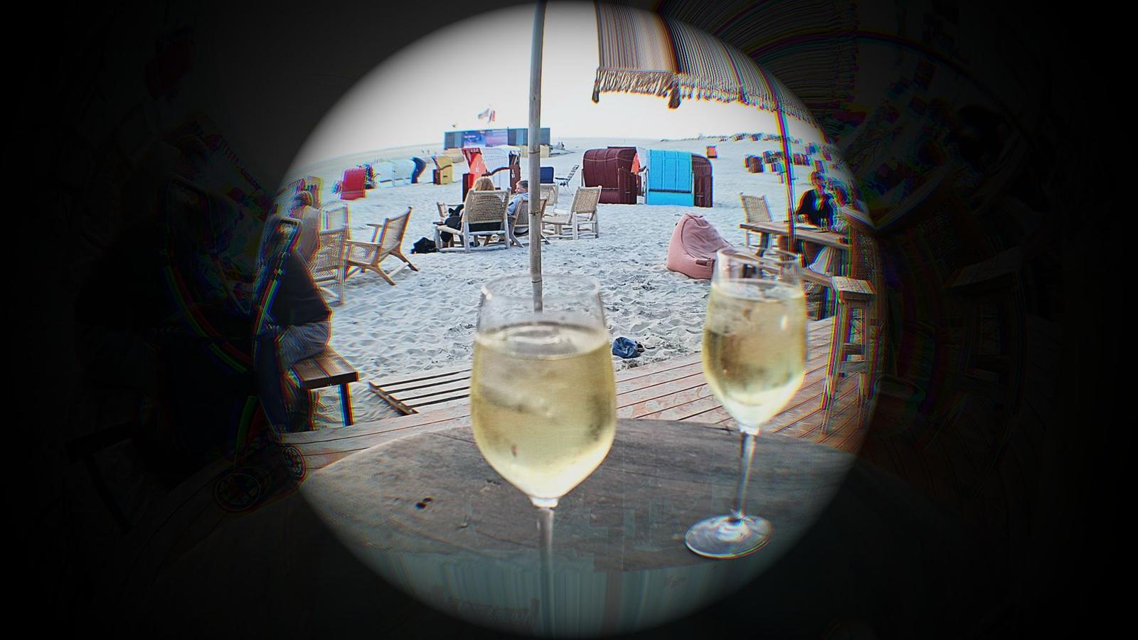 Zwei Gläser Sekt auf einem Tisch, Blick auf einen Strand mit Liegestühlen und bunten Strandkörben.