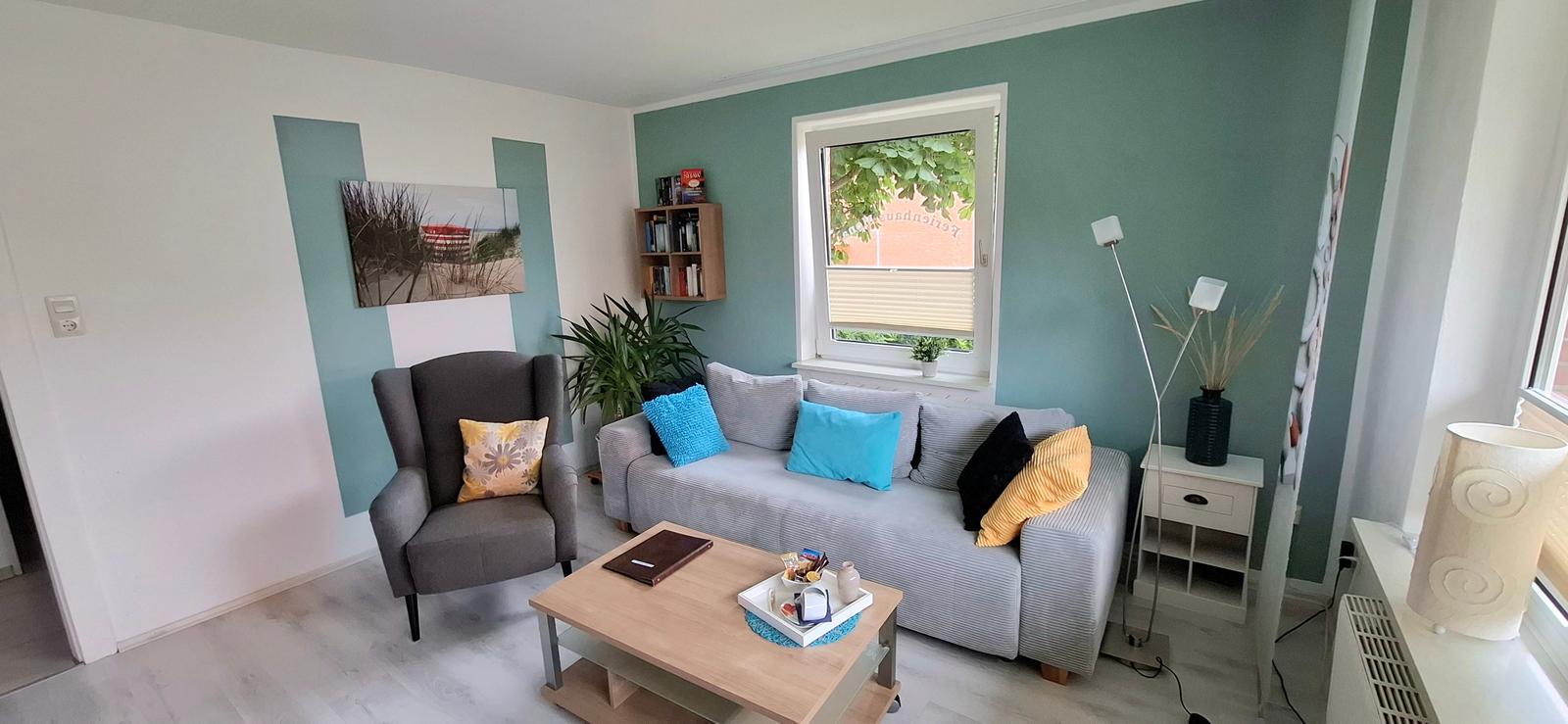 Wohnzimmer mit Sofa, Sessel und Tisch. Wandfarbe grün-weiß. Fenster mit Jalousie.