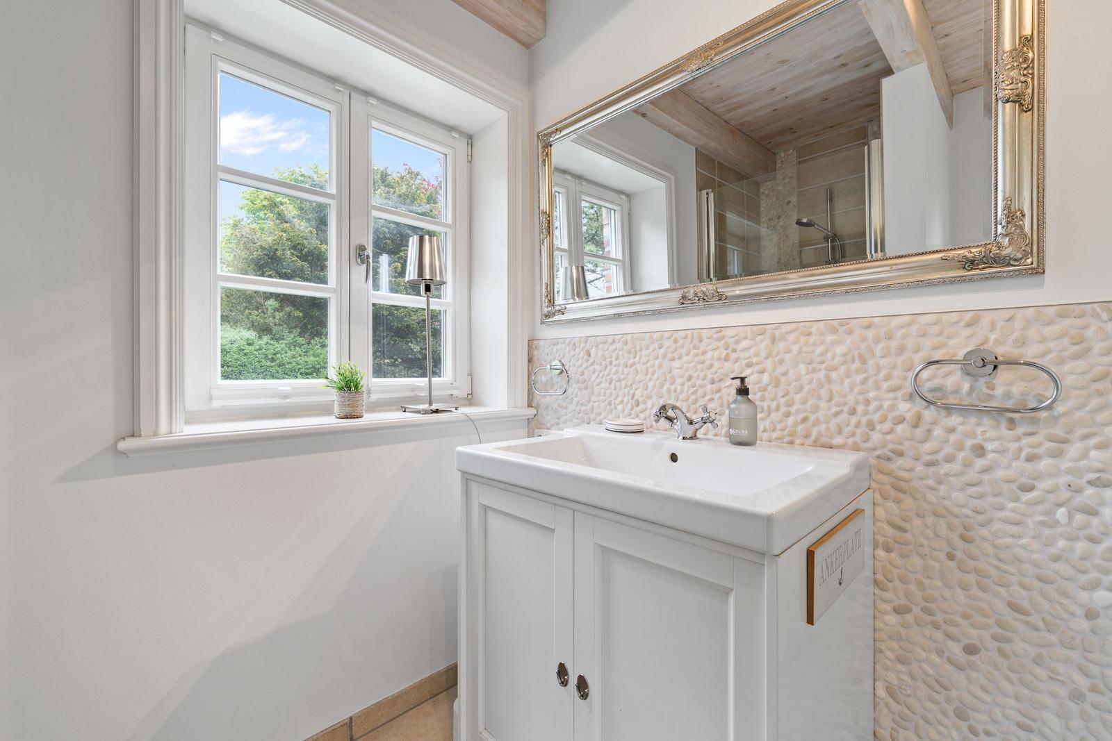 Badezimmer mit Waschbecken, Spiegel und Fenster mit Blick auf Grün.