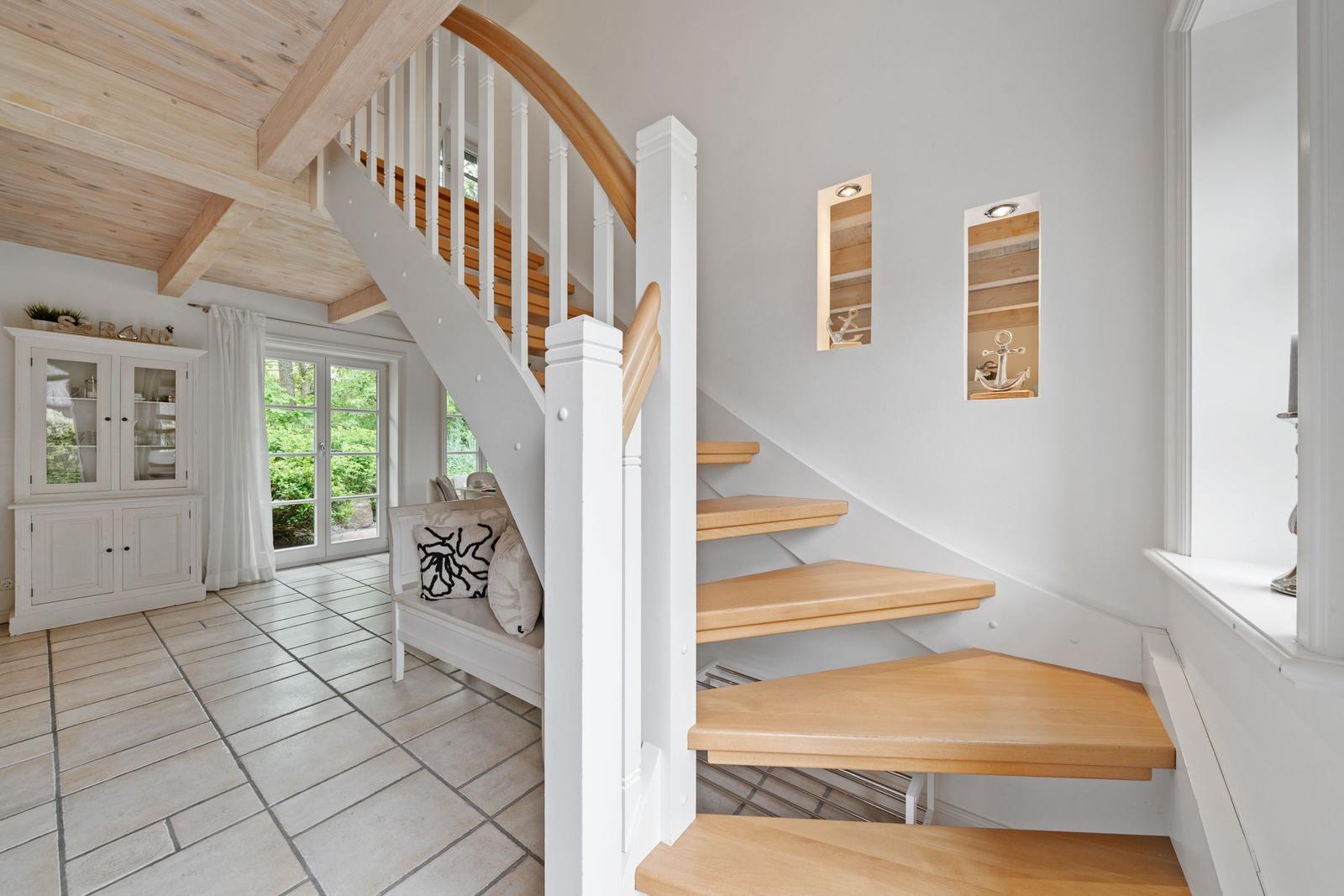 Treppenhaus mit Holzstufen und weißem Geländer. Im Hintergrund steht ein weißer Schrank und ein Fenster.