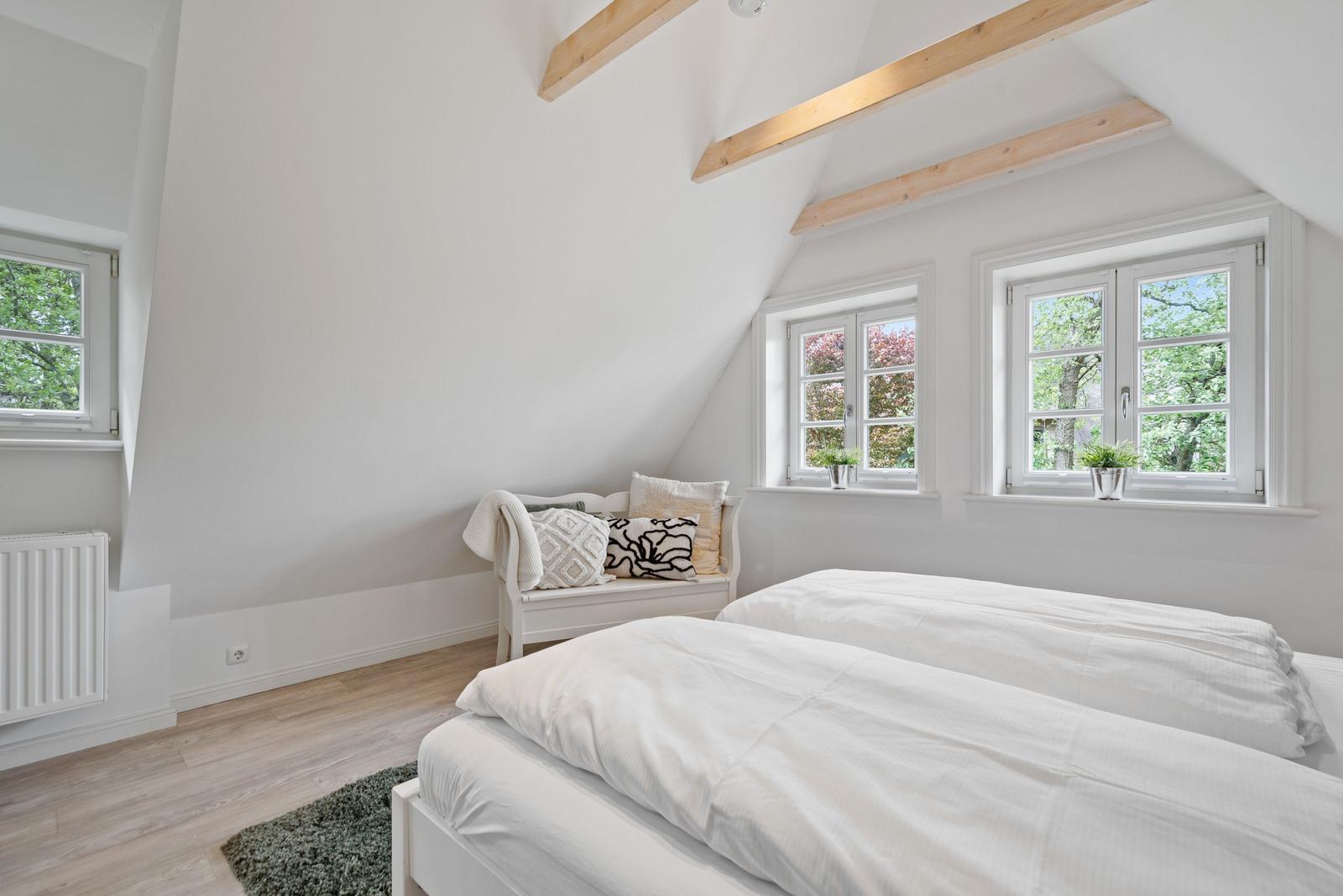 Zimmer mit zwei Betten, Holzbalken an der Decke und Fenstern mit Blick auf Bäume.