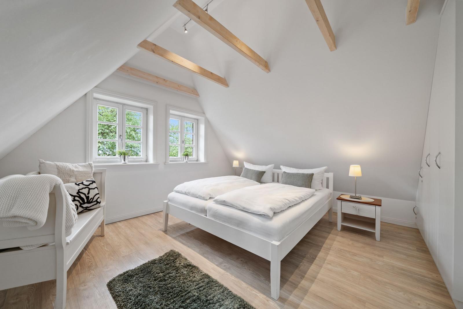 Schlafzimmer mit Dachschrägen, zwei Fenstern, Bett, Nachttisch und Schrank.