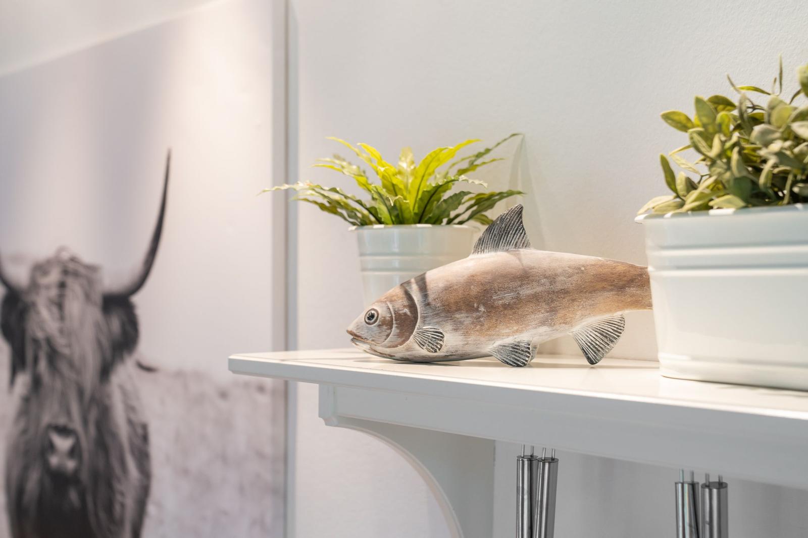 Ein Holzfigur eines Fisches steht auf einer weißen Ablagefläche neben zwei Pflanzen in weißen Töpfen.