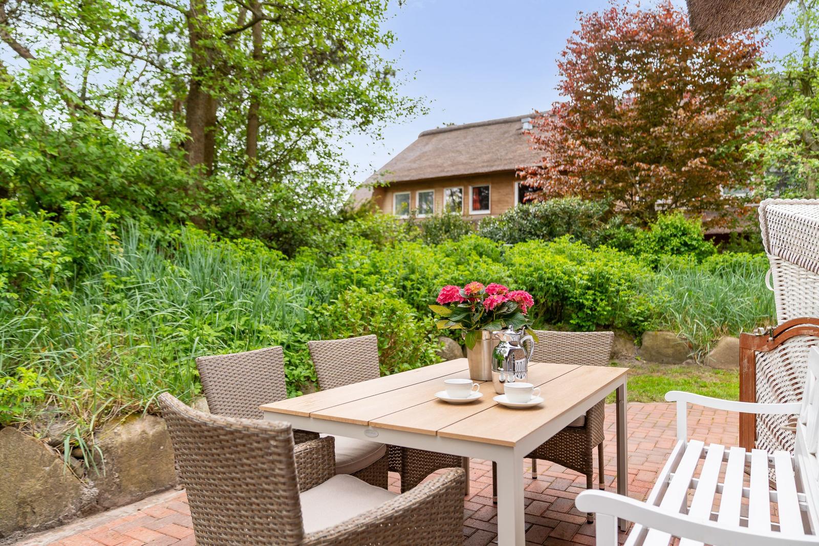 Terrasse mit Tisch, Stühlen und Blumen. Im Hintergrund ein Haus mit Schindeldach.