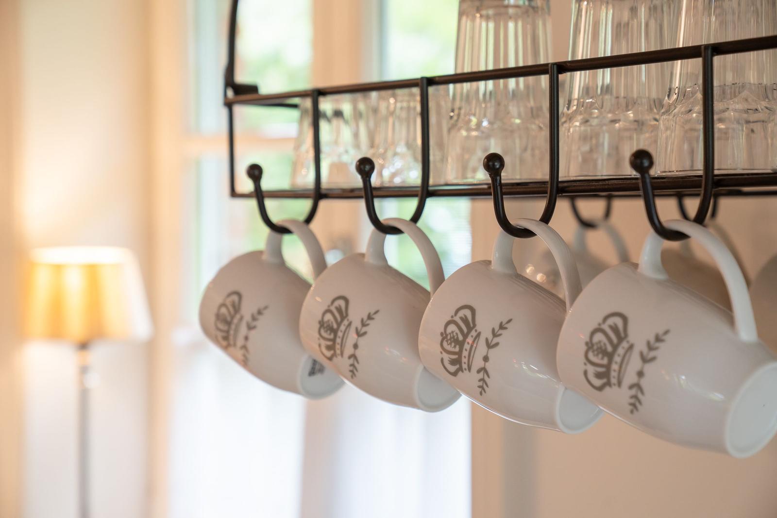 Weiße Tassen mit Krone hängen an einem schwarzen Hakenregal über Gläsern.