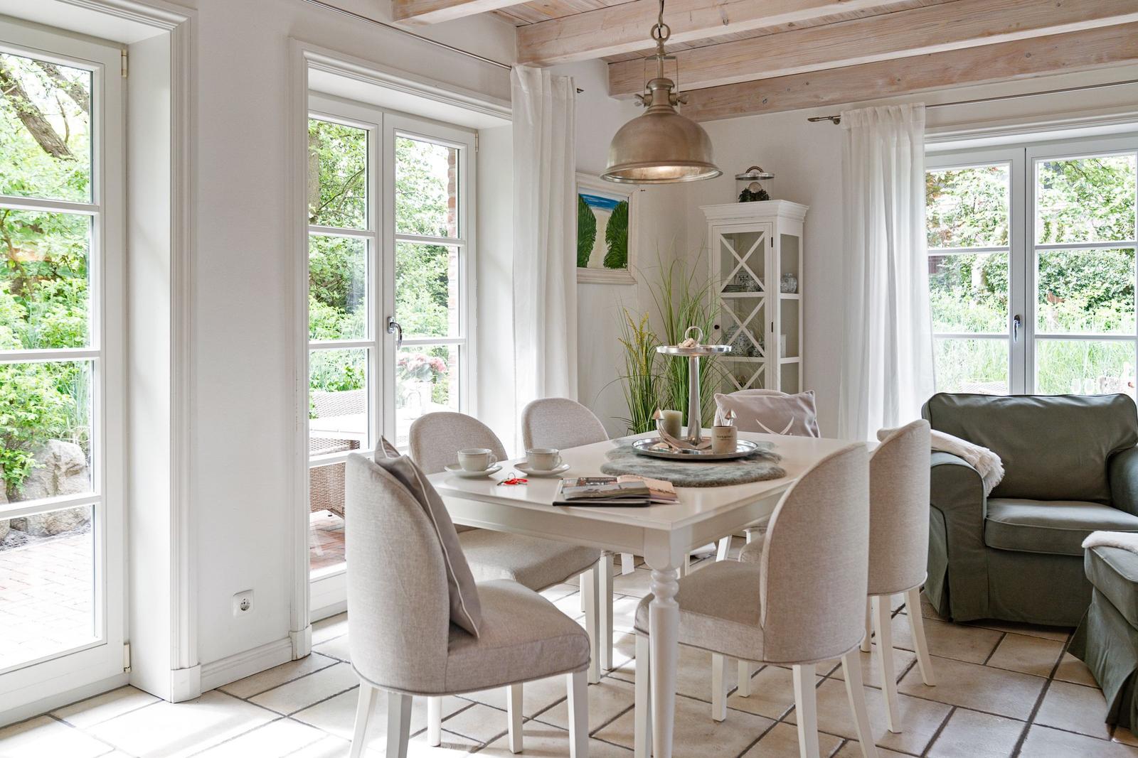 Esszimmer mit Tisch, Stühlen und Blick auf Garten. Holzbalken, Fenster und Beleuchtung sichtbar.