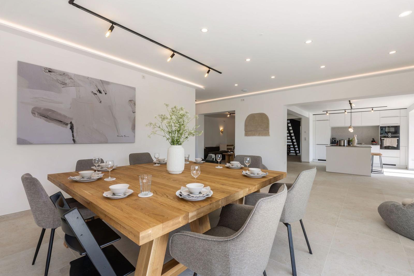 Essbereich mit Holztisch, grauen Stühlen und moderner Beleuchtung. Küche im Hintergrund.