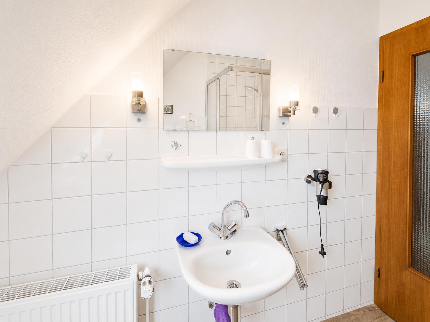 Badezimmer mit Waschbecken, Spiegel und Dusche. Weiße Fliesen und Handtuchhalter.