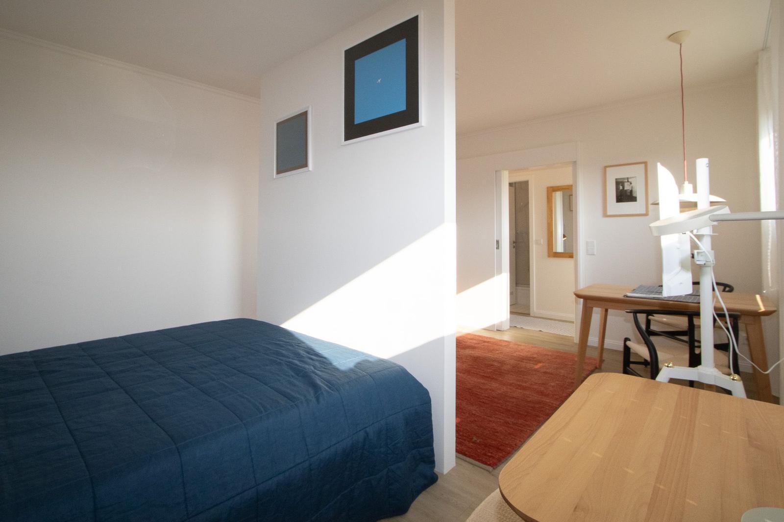Schlafzimmer mit Bett und Tisch. Blick in Wohnbereich mit Schreibtisch und Teppich.