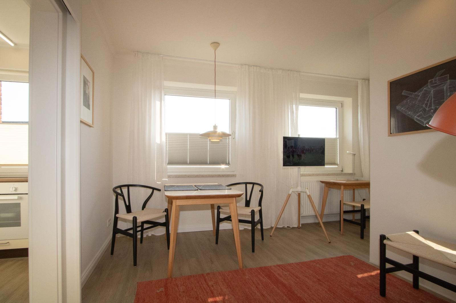 Esszimmer mit Tisch, zwei Stühlen, Fenstern und Fernseher. Holzfußboden und roter Teppich.