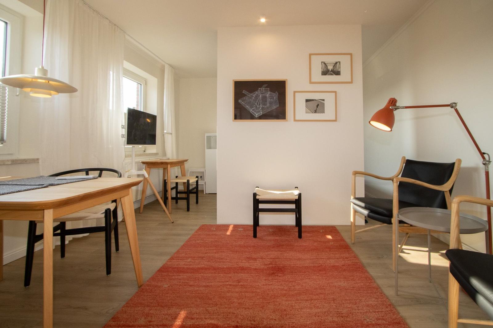Ein Raum mit Holzmöbeln, rotem Teppich und Wandbildern. Es gibt einen Schreibtisch, einen Sessel und eine Stehlampe.