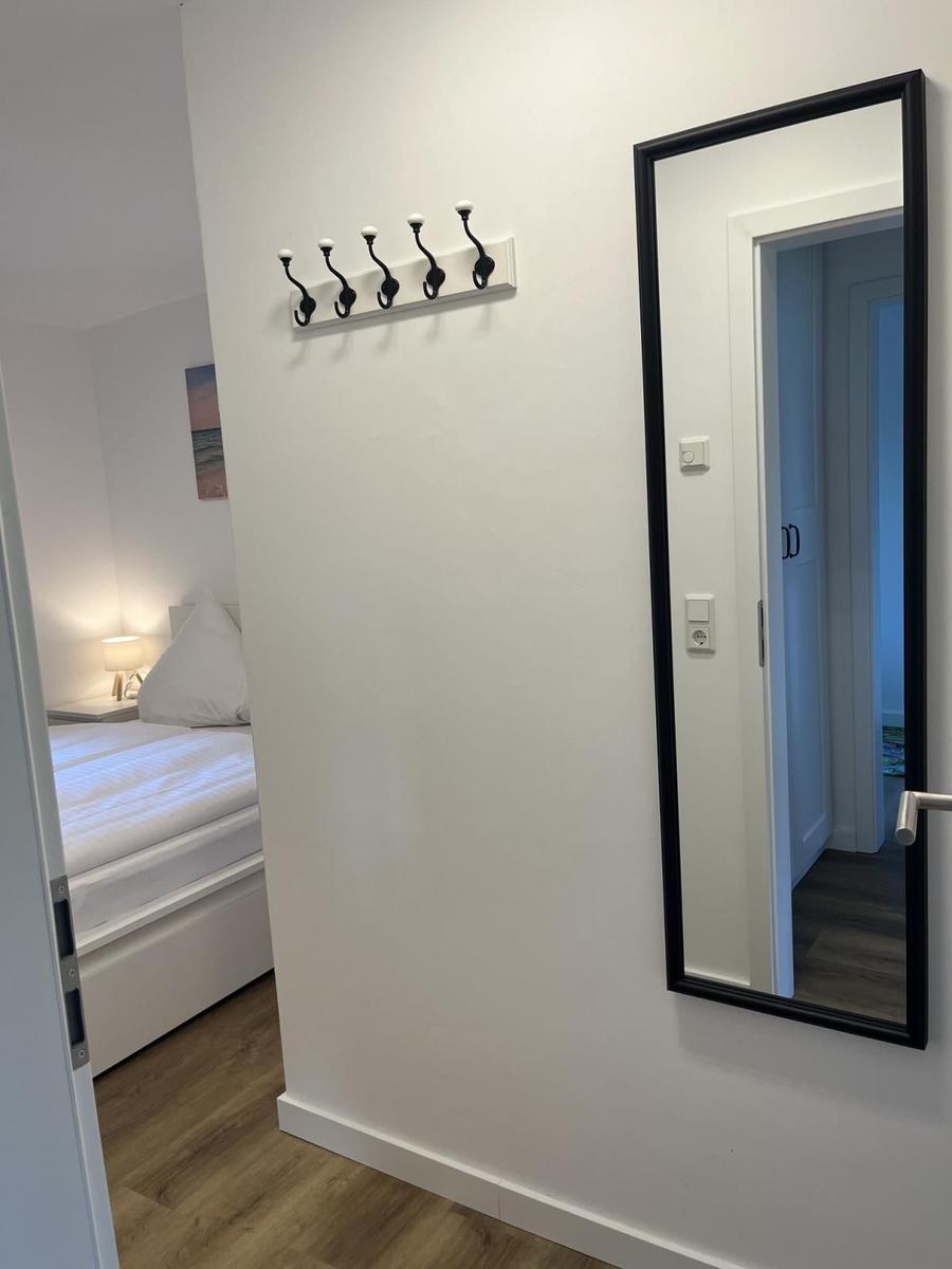 Schlafzimmer mit Bett, Wandspiegel und Hakenwand. Holzboden und weiße Wände.