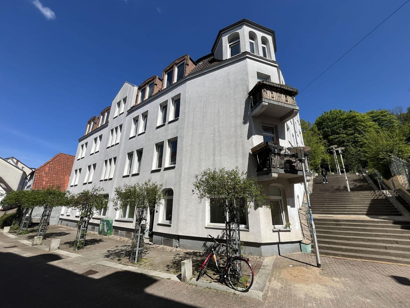 Ein mehrstöckiges Gebäude mit Balkonen und Dachfenstern, vor dem eine Straße mit Fahrrad und Bäumen verläuft.
