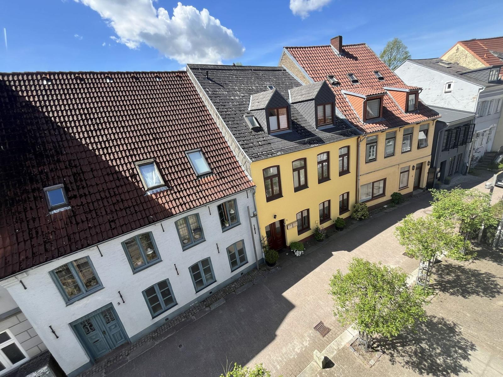 Haus mit gelber Fassade und rotem Dach in einer Straße mit gepflastertem Hof.