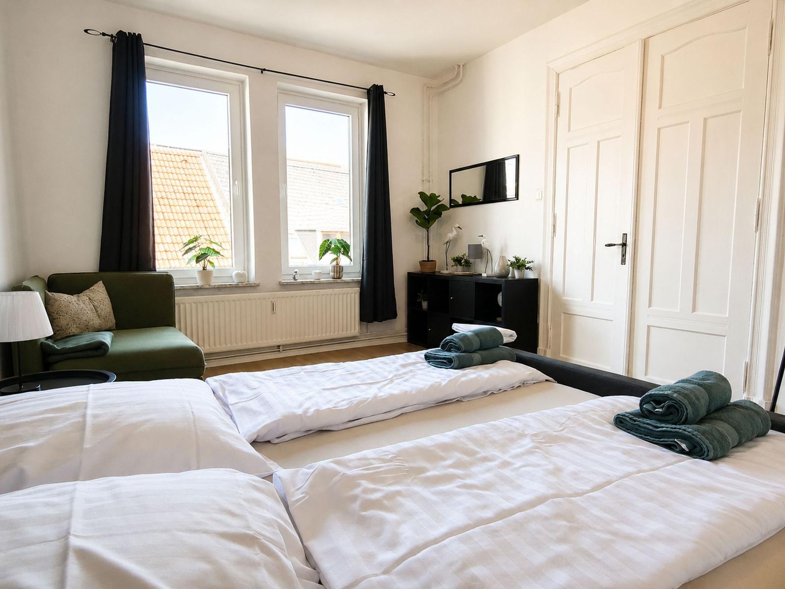 Zimmer mit zwei Betten, grünem Sofa und Fenstern mit Blick auf Dächer.